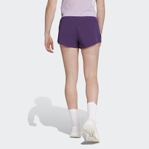 En kvinde står med ryggen til kameraet i en Adidas Adizero Gel Dame Aurplu lyslilla top, mørklilla shorts, hvide ankelsokker og hvide sko på en almindelig hvid baggrund.