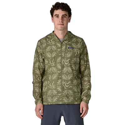 En mand står med front mod en almindelig hvid baggrund iført en Patagonia Houdini Jacket, Herre, i Sand Waves/Caper Green med hætte og logo på brystet, sammen med grå bukser.
