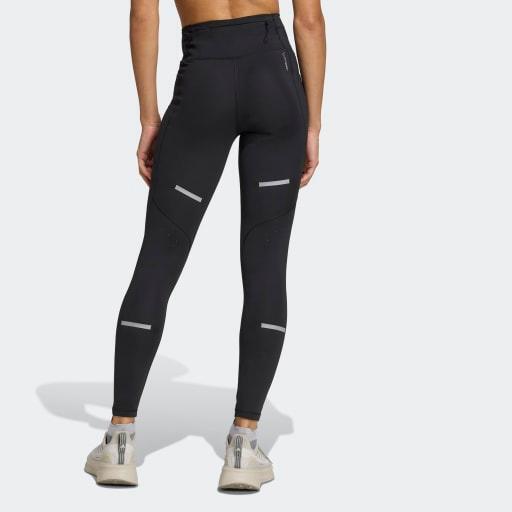 En person kigger væk og er iført Adidas Adi365 Leggins, Dame, Black med refleksstriber og grå sportssko på en almindelig lysegrå baggrund.