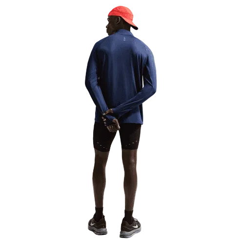 En person står med ryggen til og er iført en rød kasket, sorte shorts, sorte sportssko og en Nike Stride Half Zip Midlayer Running Top til mænd i Midnight Navy/Reflective Silver. Hænderne er foldet bag ryggen mod en lys, ensfarvet baggrund.
