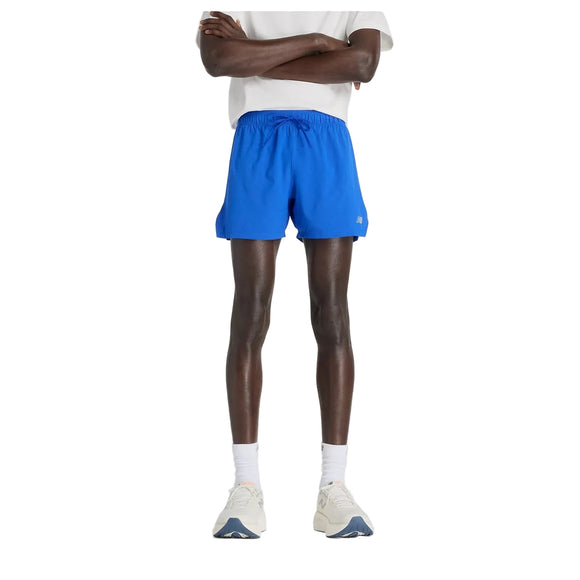 En person står på en almindelig hvid baggrund iført en hvid t-shirt, New Balance RC Short 5" Herre i Blue Gemstone, hvide strømper og hvide sneakers. Armene er krydsede, og ansigtet er ikke synligt.