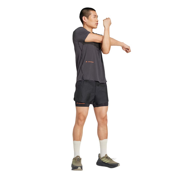 En person står på en hvid baggrund iført en Adidas Adizero x H.Koumori Running T-shirt til mænd i sort, sorte shorts, hvide sokker og løbesko, og strækker sin højre arm over brystet med sin venstre hånd.