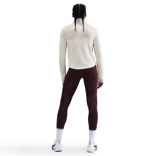 En person med lange fletninger, set bagfra mod en almindelig hvid baggrund, er iført en Nike Therma-FIT Turtle-Neck Running Top (Dame, Sail/Reflective Silv), bordeauxfarvede leggings, hvide sokker og hvide sportssko.