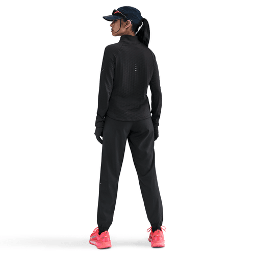 En kvinde står med ansigtet vendt mod en hvid baggrund iført en Nike Therma-FIT Turtle-Neck Running Top til kvinder i sort/reflekterende sølv. Hun har også mørke bukser, handsker, en visirhue, lyserøde sko og håret sat op i en lav hestehale.