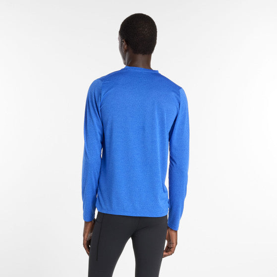 En mand kigger væk fra kameraet og er iført New Balance Athletics Long Sleeve, Herre, i Blue Gemstone Heather med sorte bukser på en almindelig hvid baggrund.