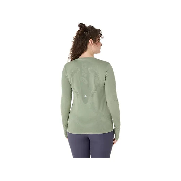 En kvinde med krøllet hår bærer en Asics Road Seamless LS Top i Monument Blue/Whisper Green og mørke bukser, her set bagfra mod en hvid baggrund. Denne åndbare træningstrøje giver både komfort og stil til træning.