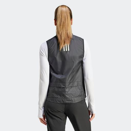 En kvinde med langt hår, set bagfra, bærer den sorte Adidas Otr B Vest Dame over en hvid langærmet skjorte og sorte bukser. Vesten har tre hvide, lodrette striber. Baggrunden er ensfarvet lysegrå. Ideel som dame løbetøj.