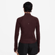 En kvinde ses bagfra iført Nike Dri-FIT 1/2-Zip Running Top, Dame, Burgundy Crush/Reflective Silv, med reflekterende detaljer og sorte shorts på en almindelig hvid baggrund. Toppen har en lille refleks på den øverste del af ryggen.