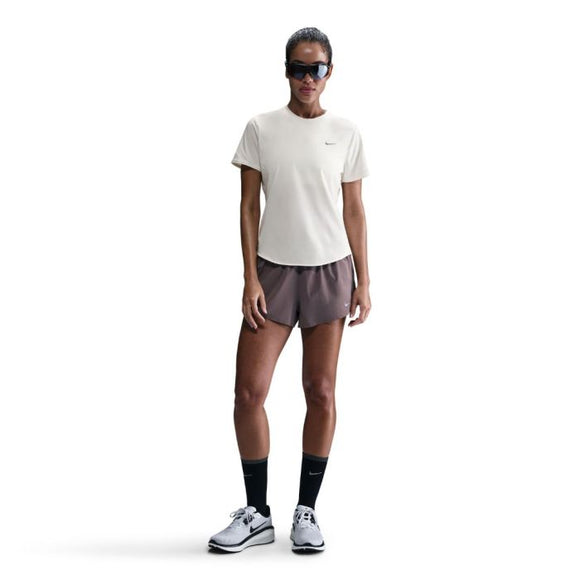 En person står og kigger fremad iført Nike Swift SS Running Top, Dame, i Sail/Reflective Silver, parret med brune sportsshorts, sorte strømper midt på læggen, hvide løbesko og mørke solbriller mod en almindelig hvid baggrund.