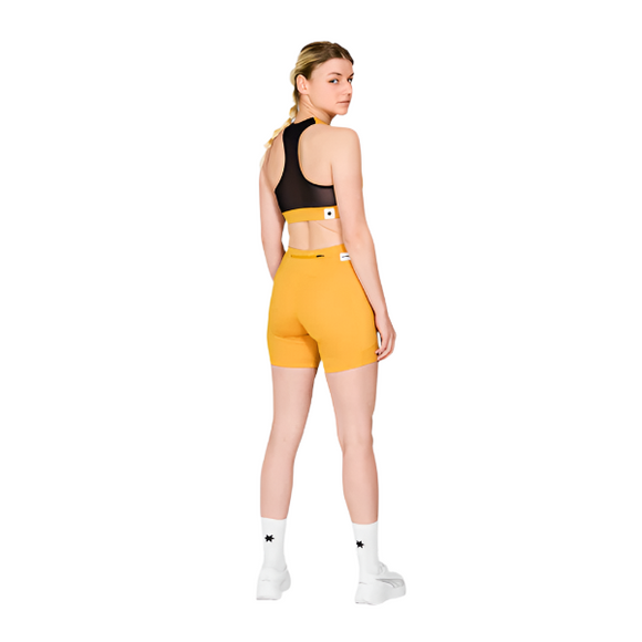 En kvinde kigger væk iført sort sports-bh, Saysky W Combat+ Short Tights 7" Dame i gul, hvide strømper og hvide sportssko på en almindelig hvid baggrund.