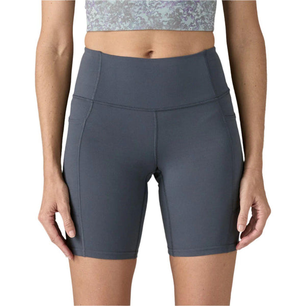 En person vises fra taljen til midt på låret iført Patagonia W's Maipo Shorts - 8 in., Dame, i Smolder Blue, sammen med en cropped mønstret top; de står med armene afslappet langs siden.