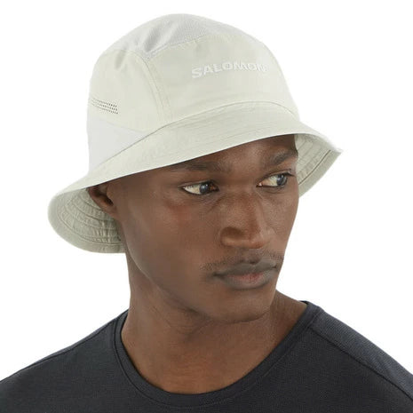 En mand bærer en Salomon SHAKEout Bucket Hat, Unisex, Icicle, med solbeskyttelse og "SALOMON" på forsiden, sammen med en mørkegrå skjorte med rund hals, og kigger lidt til siden mod en ensfarvet baggrund.