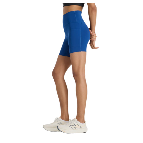En kvinde bærer en sort crop top og New Balance Sleek Pocket High Rise Short 6" i Galaxy Blue, lavet af fugttransporterende stof med sidelommer, sammen med hvide sneakers. Fra siden ses hendes overkrop og ben mod en hvid baggrund.