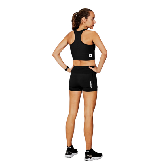 En kvinde står med front mod en sort sports-bh, Saysky W Flow+ Race Short Tights 4", Dame, Black, og sorte HOKA-løbesko. Hendes hår er bundet tilbage, og hendes hænder hviler på hofterne mod en hvid baggrund.