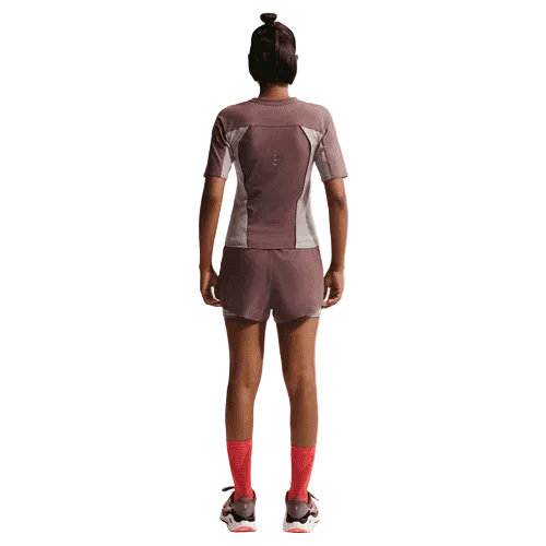 En person står med front mod kameraet iført en Nike Swift Dri-FIT kortærmet løbetop til kvinder i Tattoo/Lt Violet Ore/Reflective Silver, sammen med matchende shorts, røde sokker og sportssko på en almindelig hvid baggrund.