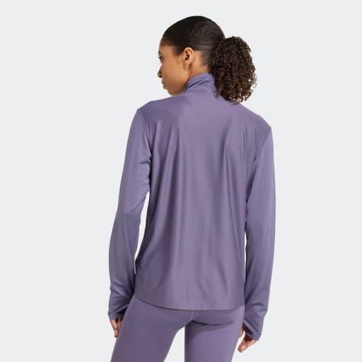 En kvinde står med ryggen til kameraet iført Adidas Otr B Hzip Dame Previo lilla træningstrøje med AEROREADY-teknologi og matchende leggings. Den langærmede, højhalsede top med halv lynlås står i kontrast til den ensfarvede lysegrå baggrund.