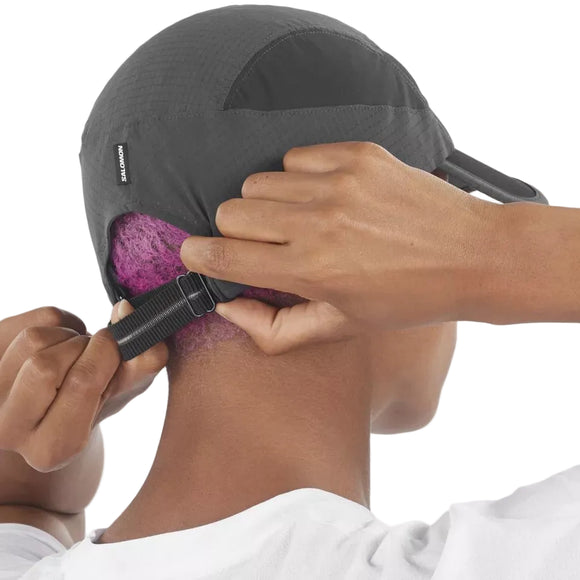 En person med kort, lilla hår justerer remmen på en Salomon Sense Aero Cap, Unisex, Deep Black. Vedkommende har en hvid skjorte på mod en almindelig hvid baggrund.
