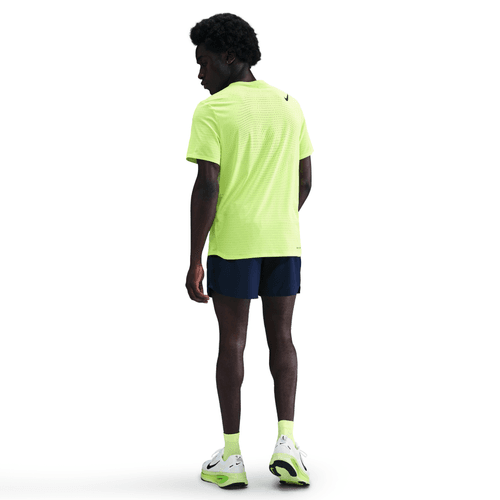 En person står med ryggen til og er iført en lys Volt/Sort Nike Dri-FIT ADV kortærmet løbetop til mænd, marineblå shorts, neongule sokker og hvide sportssko med neonfarvede detaljer på en almindelig hvid baggrund.