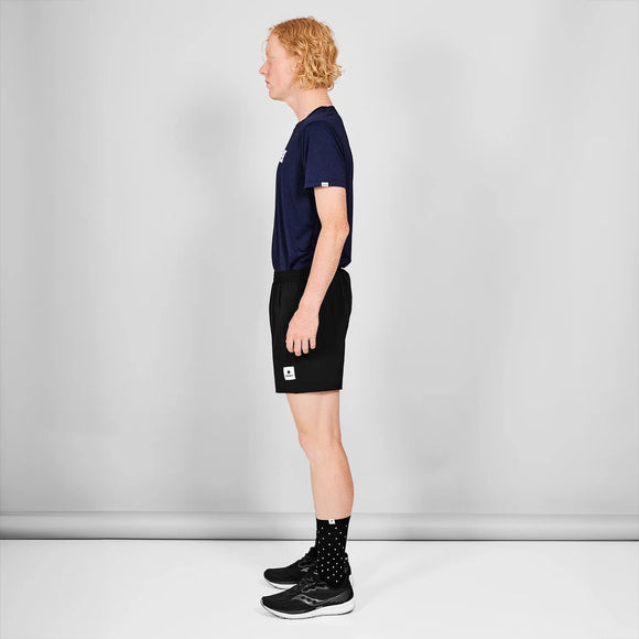 En person med krøllet blondt hår står i profil mod en almindelig hvid baggrund iført en marineblå T-shirt, Saysky Pace Shorts 5" Herre Black, sorte sokker med hvide prikker og sorte sneakers.