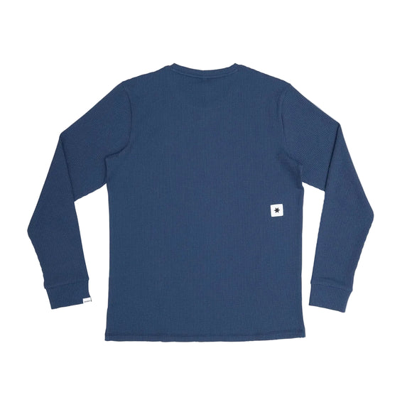 En Saysky Motion Waffle Long Sleeve, Herre, i blå ligger fladt med ryggen opad. Et lille hvidt rektangulært mærke med et sort stjernelignende symbol fremhæver den nederste sidekant på denne tekniske top med struktur.