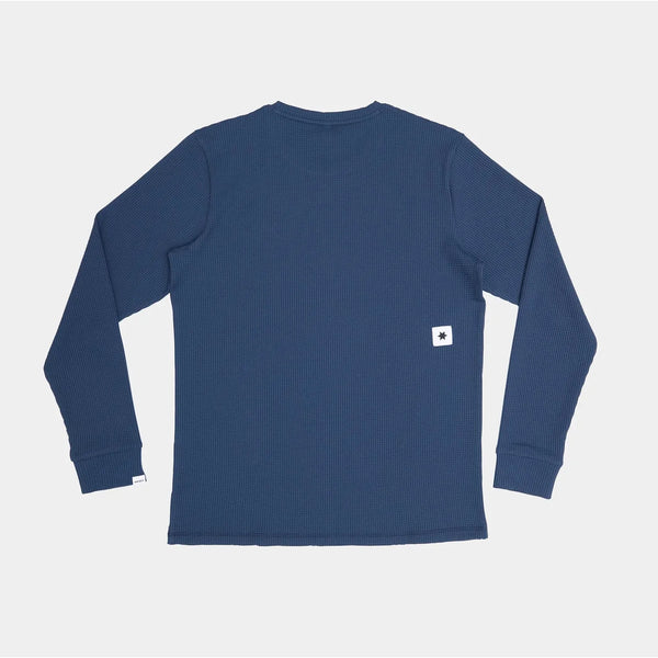 En Saysky Motion Waffle Long Sleeve, Herre, i blå ligger fladt med ryggen opad. Et lille hvidt rektangulært mærke med et sort stjernelignende symbol fremhæver den nederste sidekant på denne tekniske top med struktur.