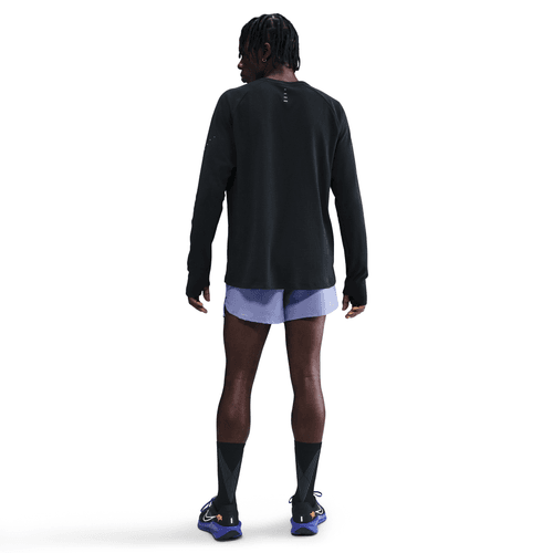 En person kigger væk fra kameraet i en Nike Dri-FIT Running Waffle Crew, Herre, Black/Reflective Silv, sammen med lyslilla shorts, sorte knæstrømper og blå løbesko på en hvid baggrund.