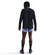 En person kigger væk fra kameraet i en Nike Dri-FIT Running Waffle Crew, Herre, Black/Reflective Silv, sammen med lyslilla shorts, sorte knæstrømper og blå løbesko på en hvid baggrund.