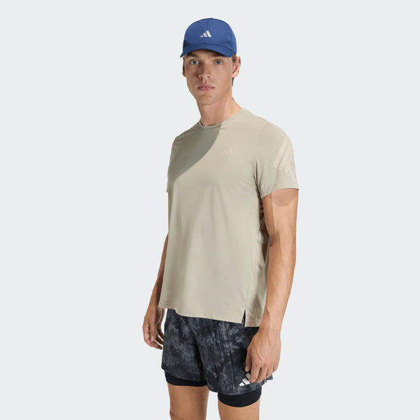 En mand er iført en Adidas Adi365 Running Iconic T-Shirt, Herre, Woca i beige, sorte og grå mønstrede shorts og en blå adidas-kasket med hvidt logo. Han står med ansigtet fremad mod en almindelig lys baggrund.
