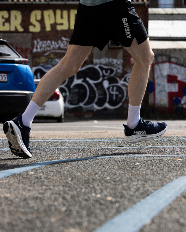 En person iført sorte shorts, hvide sokker og marineblå Hoka-løbesko jogger eller løber på en udendørs bane. I baggrunden ses biler og graffiti på en murstensbygning.