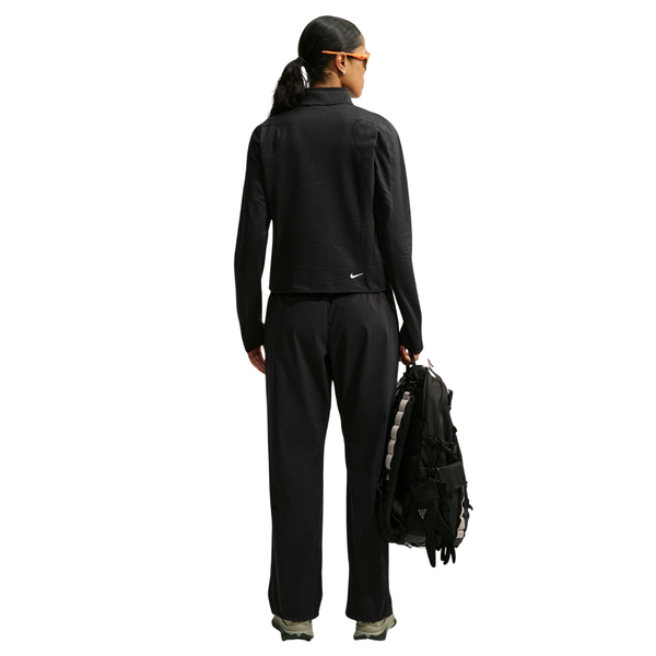 En person står med ryggen til, iført en sort Nike Therma-FIT 1/2-Zip Top og sorte bukser, og holder en sort rygsæk.