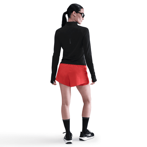 En kvinde i Nike Dri-FIT Full-Zip Running Mid Layer for dame (sort/reflekterende sølv) og røde shorts poserer med sorte sokker, sportssko, solbriller og en hestehale på en almindelig hvid baggrund.