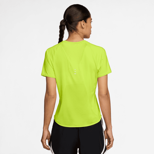 En kvinde med en enkelt fletning ses bagfra iført en Nike Dri-FIT kortærmet løbetop til kvinder i Cyber/Reflective Silver og sorte shorts med hvide striber, stående på en almindelig hvid baggrund.