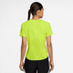 En kvinde med en enkelt fletning ses bagfra iført en Nike Dri-FIT kortærmet løbetop til kvinder i Cyber/Reflective Silver og sorte shorts med hvide striber, stående på en almindelig hvid baggrund.
