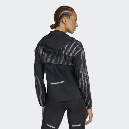 En person med flettet hår iført Adidas Adi365 Jacket, Dame, i Black/Refsil med hætte og matchende sorte leggings, står med ryggen til og kigger til siden mod en almindelig lys baggrund.