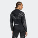 En person med flettet hår iført Adidas Adi365 Jacket, Dame, i Black/Refsil med hætte og matchende sorte leggings, står med ryggen til og kigger til siden mod en almindelig lys baggrund.