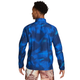 En person står med ryggen til og har en Nike Miler UV Repel-løbejakke til mænd i farven Midnight Navy/Reflective Silver på samt flerfarvede shorts med et abstrakt mønster. Baggrunden er ensfarvet hvid.