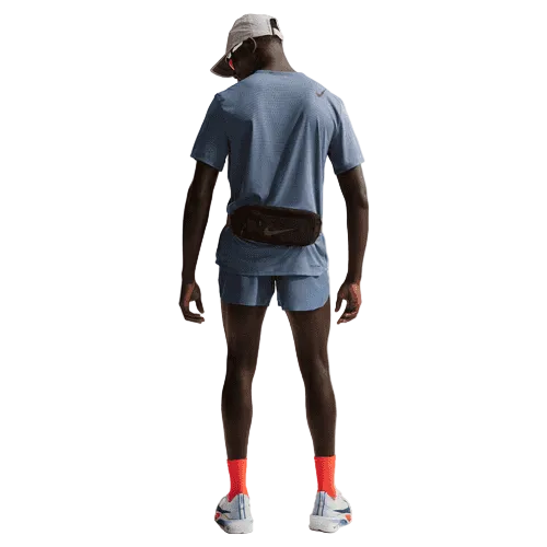 En person kigger væk, klædt i en lyseblå sportstrøje, Nike AeroSwift 4In Brief-Lined Running Shorts for men i Diffused Blue/Black, en lysegrå kasket, orange sokker, hvide løbesko og en sort bæltetaske. Baggrunden er almindelig hvid.