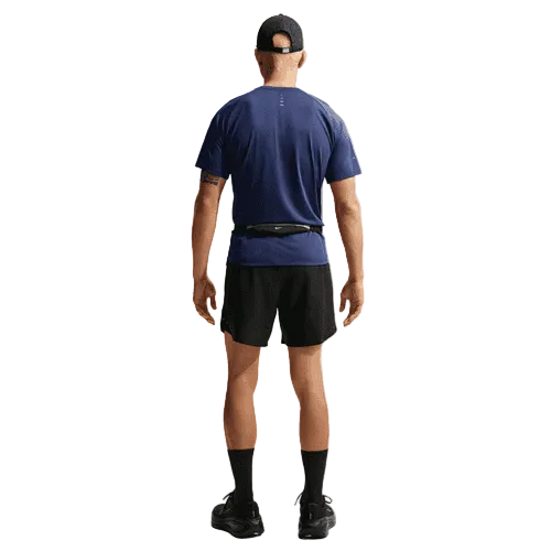 En person ser væk og er iført en Nike Stride SS Running Top Herre i Midnight Navy/Reflective Silv, sorte shorts, strømper, sko, en sort kasket og en smal sort taljetaske på en almindelig hvid baggrund.