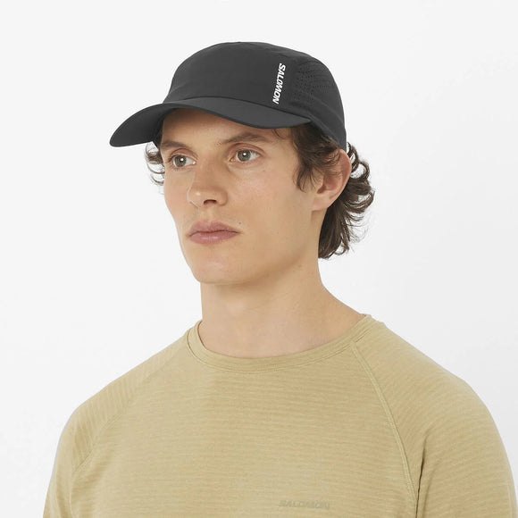 En person i en beige skjorte og en Salomon SHKOUT Cap U, Deep Black, med justerbar pasform ser lidt skævt ud mod en almindelig hvid baggrund.