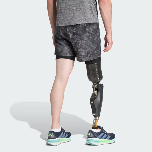 En person iført grå skjorte, Adidas Otr Sd 2In1 M Herre løbeshorts i Grefou/Black og blå sneakers står med ansigtet vendt væk, og en protese på højre ben er synlig under knæet.