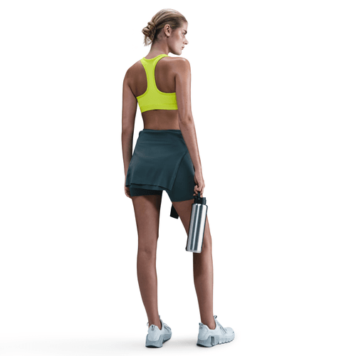 En kvinde kigger væk, iført Nike Swoosh Medium Sports Bra til kvinder i Cyber/White, sammen med en mørk atletisk nederdel, sweatshirt bundet i taljen, hvide sneakers og med en vandflaske i rustfrit stål i hånden mod en hvid baggrund.