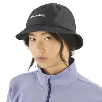 En person bærer en Salomon SHAKEout Bucket Hat, Unisex, Deep Black, og en lyslilla jakke med lynlås, mens han kigger mod kameraet på en almindelig lys baggrund.
