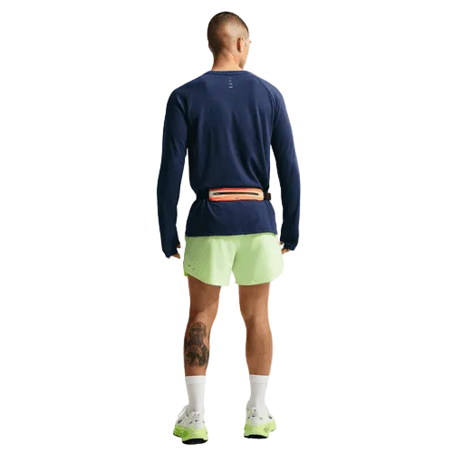 En person vender ansigtet væk, iført en Nike Stride Waffle Crew løbetrøje til mænd i Midnight Navy/Reflective Silver, lysegrønne shorts, hvide sokker, neongrønne løbesko, med refleksdetaljer og en lille taske spændt fast i taljen.