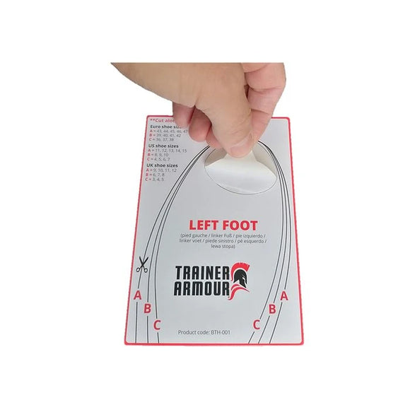 En hånd holder en Trainer Armour Big Toe Hole Preventer-størrelsesguide (Unisex, hvid) mærket LEFT FOOT med størrelsesinformation, snitlinjer (A, B, C), en cirkulær fodudskæring og produktkoden BTH-001 nederst.