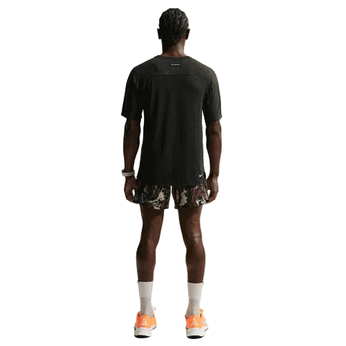 En person med flettet hår kigger væk fra kameraet iført en sort Nike ACG "Solar Chase" Trail Running Top (Herre, Black/Black/Summit White), camouflageshorts, hvide sokker og orange sportssko på en hvid baggrund.