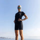 En person står udendørs i en LiiteGuard Ground-Tech T-shirt, Dame, Black, sammen med shorts og reflekterende solbriller. Den lyse himmel og den fjerne kystlinje skaber en naturskøn baggrund.