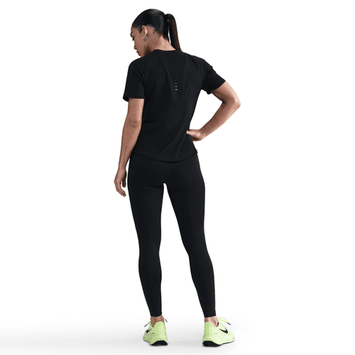 En person med hestehale er iført en sort t-shirt, Nike Therma-FIT ADV High-Waisted 7/8 Reflective Running Leggings (Dame, Black/Dk Smoke Grey) og neongrønne sportssko. Højre hånd hviler på hoften mod en almindelig hvid baggrund.