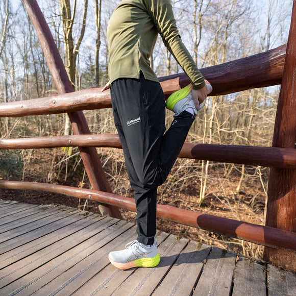 En person iført Liiteguard Re-Liite Long Pants, Dame, Black strækker sit højre ben ved at holde foden bag sig, mens han står på en træbro omgivet af træer.