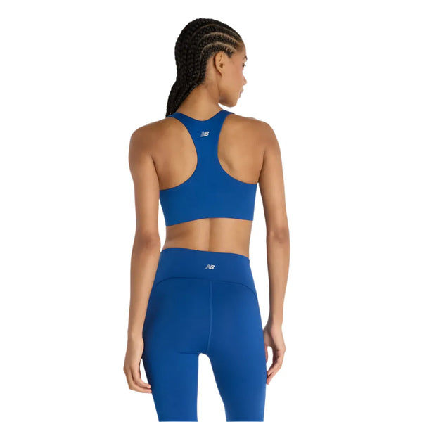 En kvinde med flettet hår ses bagfra iført New Balance Define Train Bra, Dame, i Galaxy Blue, med matchende leggings og synlige logoer, stående på en almindelig hvid baggrund.