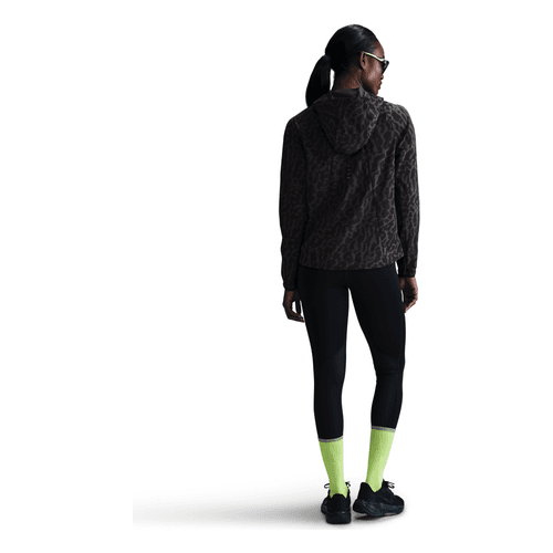 En person i Nike Dri-FIT løbejakke til kvinder (Sort/Anthracite/Reflective Silver), sorte leggings, gulgrønne sokker, sorte sneakers og solbriller står med hovedet vendt lidt til venstre.